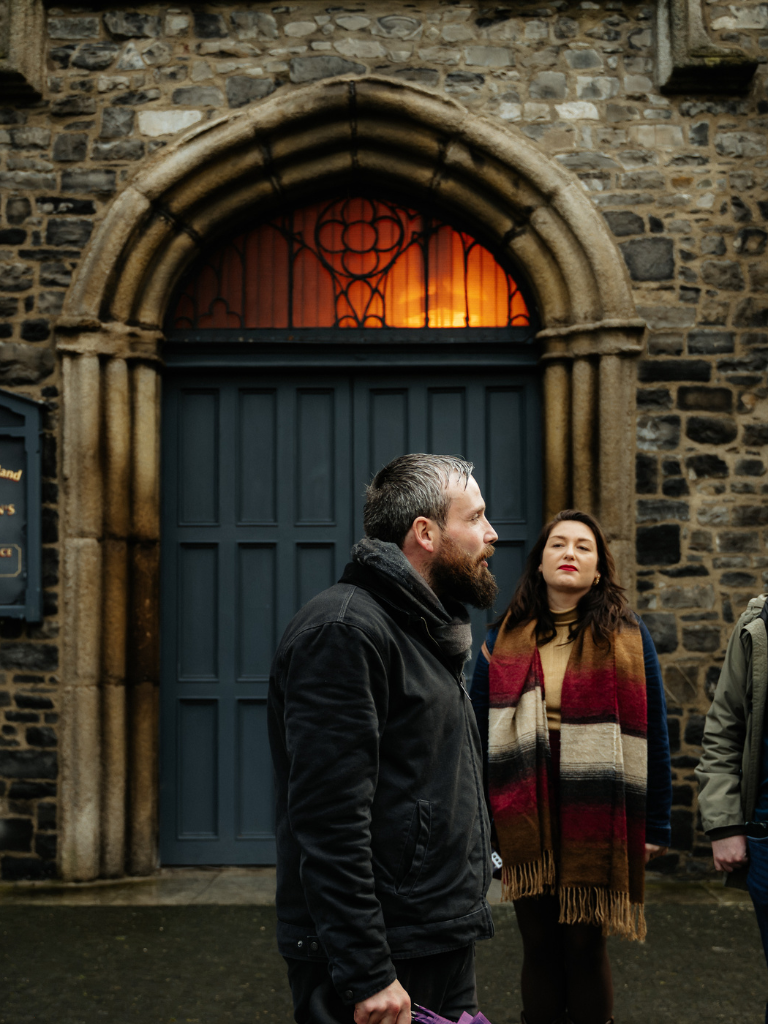 Unearthed Tours guide leading the Dark Dublin Tour to a group of tourists in front of a historic church and graveyard at dusk.